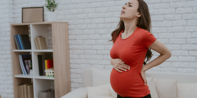 امرأة حامل تقف واضعة يدها اليسرى خلف ظهرها و يدها اليمنى اعلى بطنها وهي ترفع رأسها متألمة  