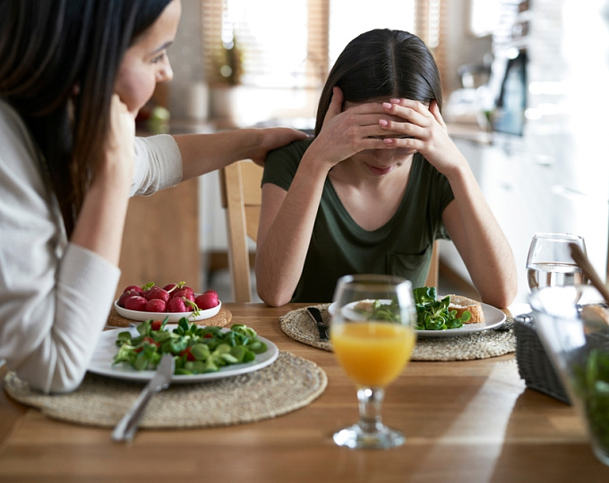 ام تضع يدها على كتف ابنتها تدعمها لتناول طعامها و الفتاة تضع يديها على رأسها وامامها طبق من الطعام و كأس عصير 