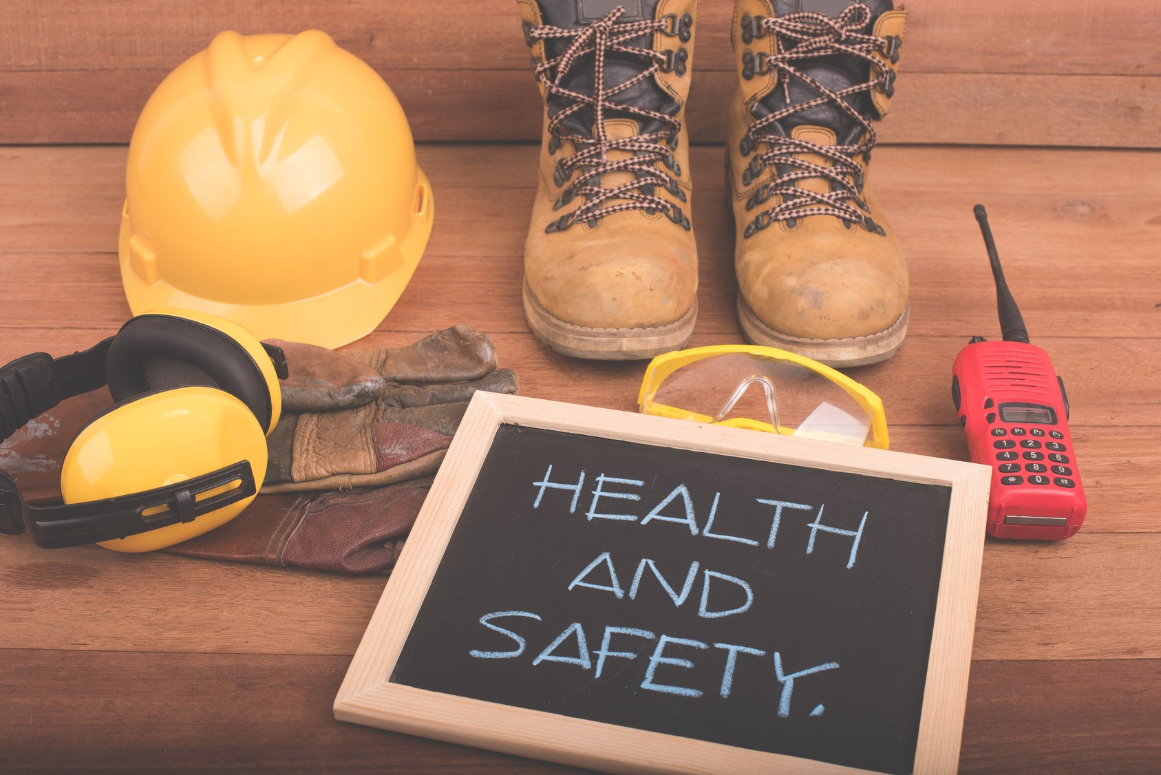 على ارضية خشبية تظهر معدات سلامة للعمل تشمل خوذة صفراء و سماعات لحماية الاذن و حذاء بني و نظارات و لجهاز اتصال لاسلكي و لوح حشبي مكتوب عليه HEALTH AND SAFETY