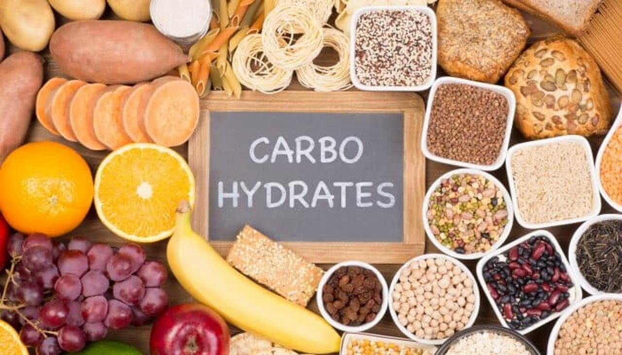 لوح اسود صغير مكتوب عليه كلمة CARBO HYDRATES  وحوله مجموعة كبيرة من الاغذية الغنية بالكربوهيدات مثل الليمون والموز البطاطا الحلوة و العنب والتفاح الاحمر و والزبيب والحمص والمعكرونة و الخبز والفاصولياء 