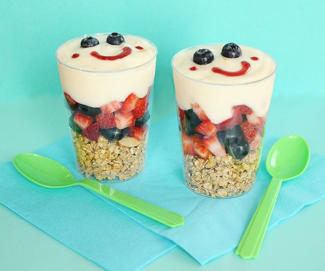 كوبين شفافين من بارفيه الزبادي وتظهر من خلاله طبقات الفواكه ومرسوم على سطحه وجه بالفواكهة وبجانب الكاسين ملعقتين خضراوين 