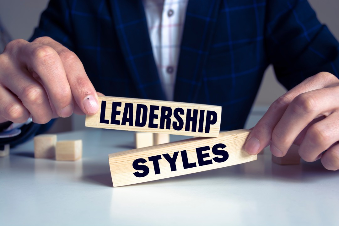 القيادة شخص يجلس الى طاولة ويمسك بلوحين مكتوب عليهما LEADERSHI[ STYLES
