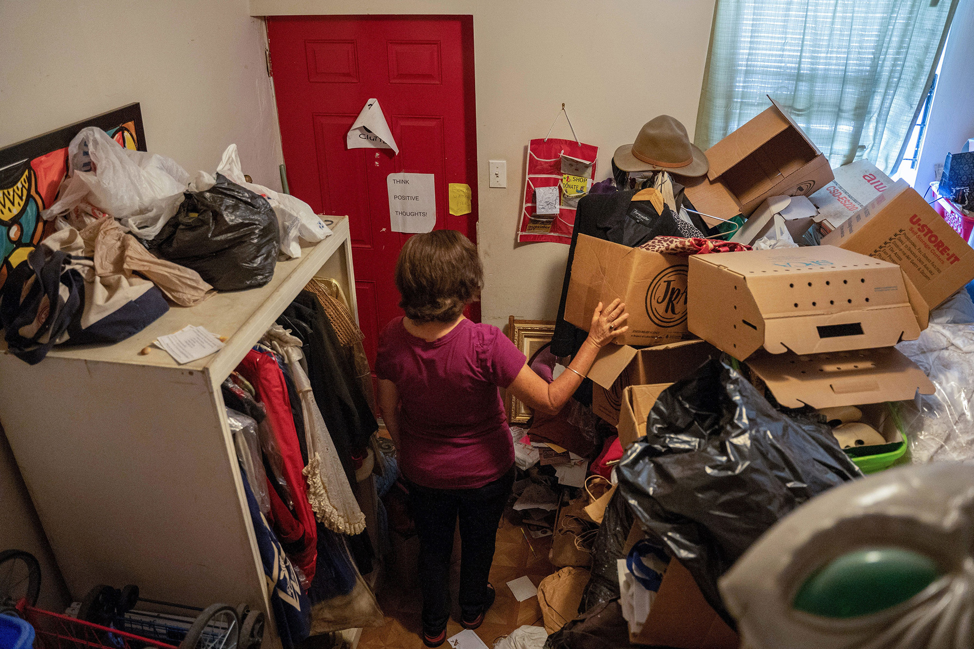 منزل تتكدس فيه الاغراض من صناديق و ملابس و اكياس و سيدة تقف في وسط الغرفة 