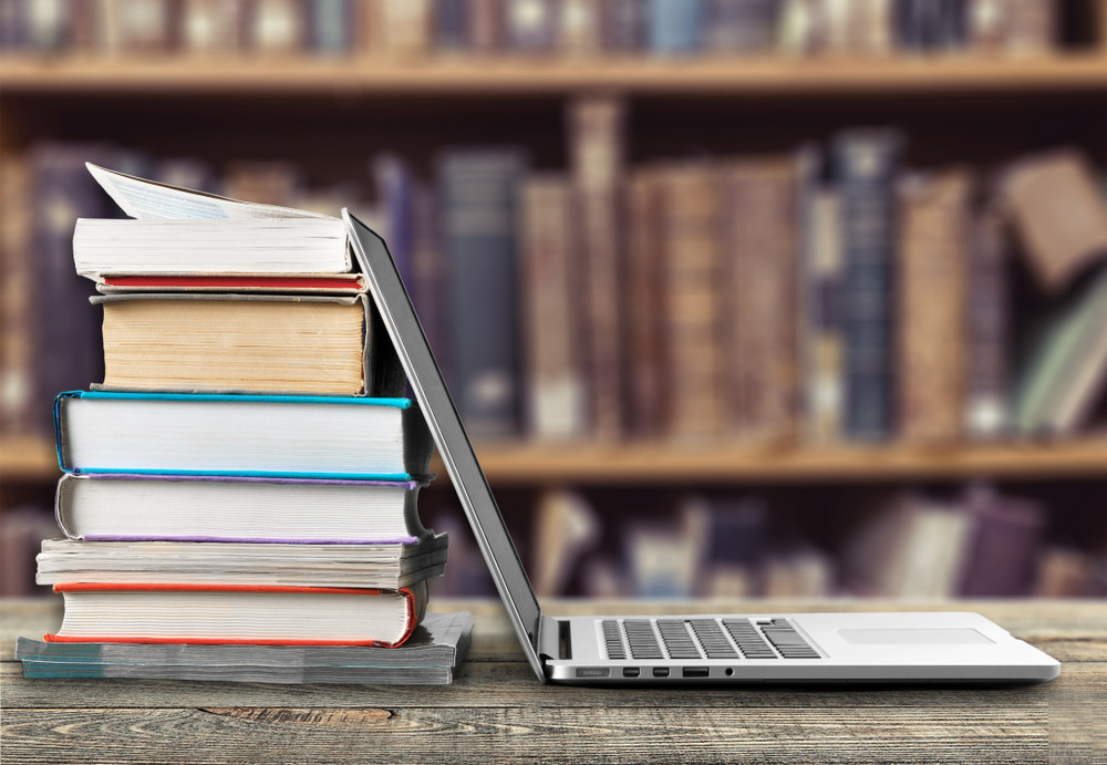 ضمن مكتبة يظهر على الطاولة مجموعة من الكتب و بجابنها لابتوب مفتوح 