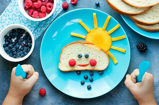 طفل امامه طبق ازرق عليه قطعة من الخبز مرسومة بشكل وجهة بالفواكهة و فوقها شمس مصنوعة من الطعام 