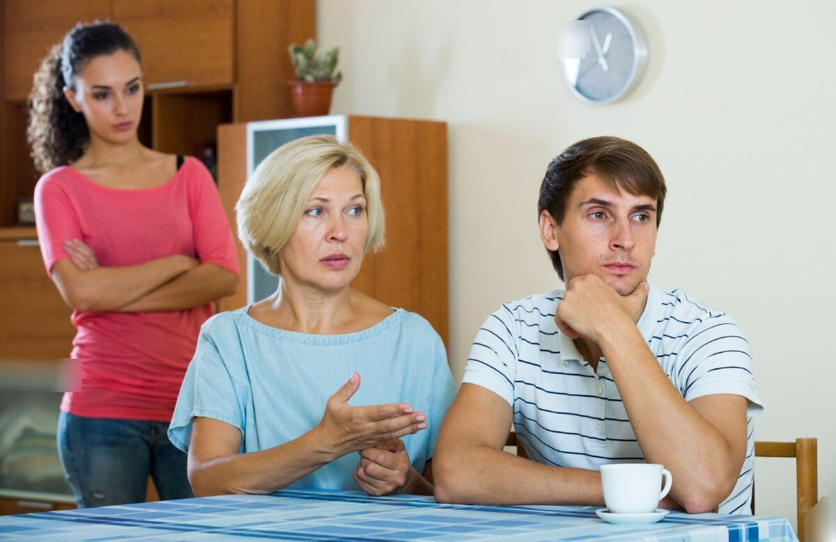 ام تتكلم مع ابنها وتتدخل في حياته مع زوجته والابن غير سعيد بهذا التدخل والزوجة تقف خلف والدته شابكة يديها الى صدرها 