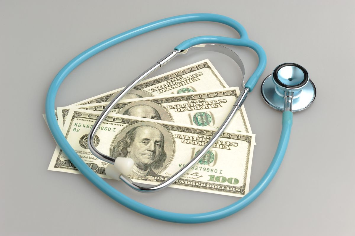 على خلفية رمادية تظهر سماعة دكتور لونها ازرق موضوعة على ثلاث اوراق مالية 