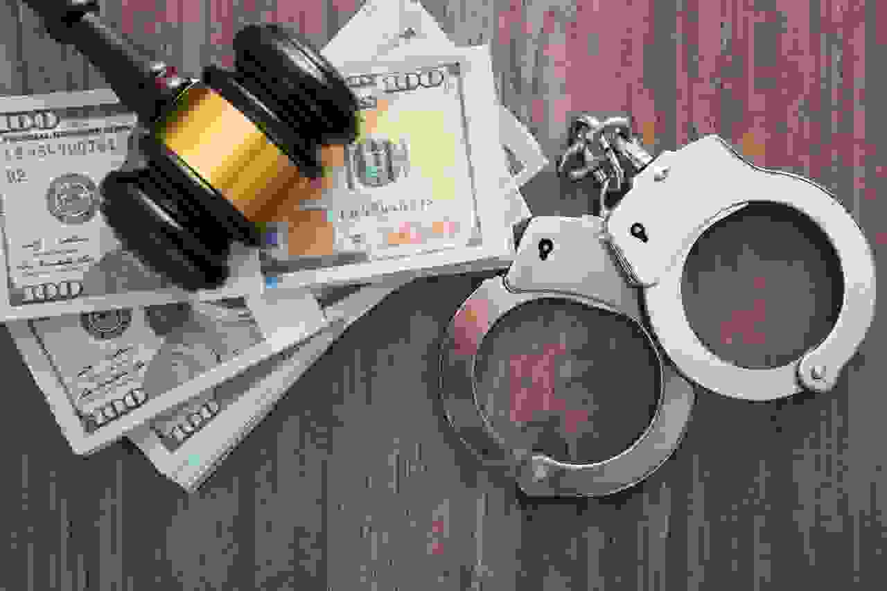 على طاولة خشبية تظهر 3 مجموعات من الاوراق المالية و فوقها مطرقة القضاء و بجانبها اصفاد السجن 