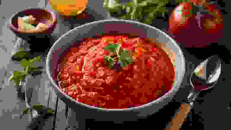 طبق يحوي صلصة المارينارا المصنوعة من البندورة مزينة بورقة بقدونس ويوجد على الطالولة حبة بندورة و صبق صغير فيه ثوم وملعقة 