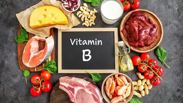 فيتامينات ب المركبة لوح خشبي مكتوب عليه VITAMIN B وحوله مجموعة من الاغذية الغنية بفيتامينات ب المركبة مثل الكبده والسمك واللحم الاحمر و الكاجو والبندورة و زيت الزيتون