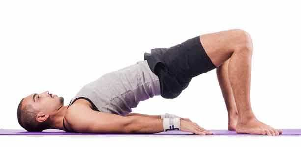 شاب يرتدي تيشير رمادي و شورت اسود يتمدد على سجادة الرياضية و يرفع االحوض للأعلى و قدميه بشكل عامودي  مثبتا يديه على الارض 