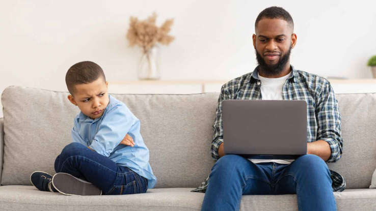 أب يجلس على الاريكة و يعمل على اللابتوب و يجلس ابنه المراق بجانبه مديرا ظهره حزينا منزعجا  