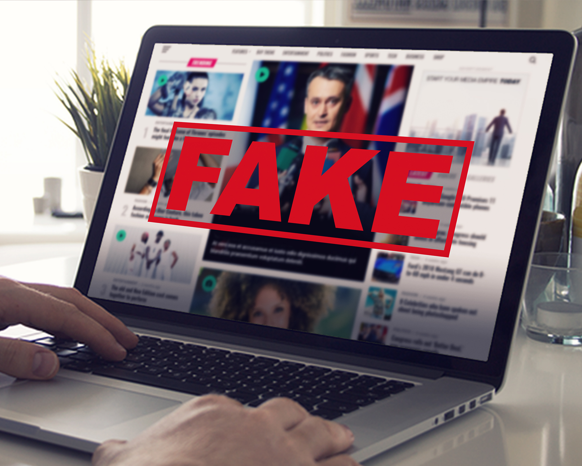 اخبار مزيفة على طاولة بيضاء يظهر لابتوب ويدين تنقران على لوحة المفاتيح و يظهر على الشاشة وعليها عبارة FAKE
