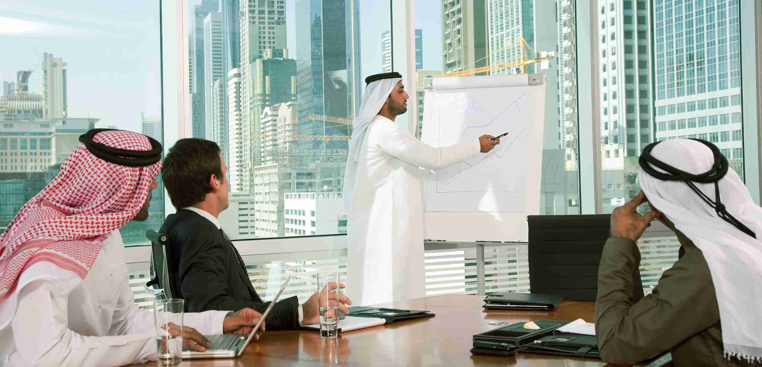 اجتماع عمل لمشروع تجاري خليجي و يقوم احد الاشخاص بشرح المشروع للمجتمعين ويطلون على منظر لابنية شاهقةخلف زجاج غرفة الاجتماع 
