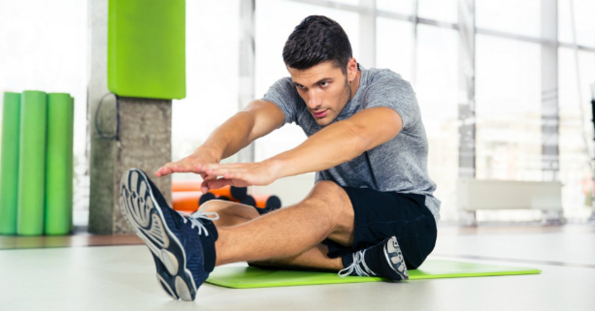 شاب في النادي الرياضي يمارس تمارين اللياقة جالسة على سجادة التمرين و يمد يديه ليلامس قدمه المفروده 