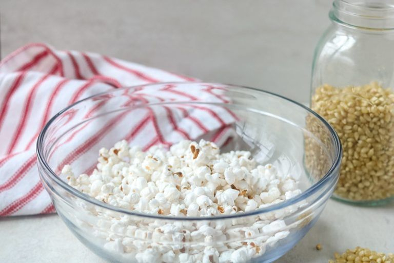 على طاولة بيضاء يظهر وعاء زجاجي شفاف بداخله فشار و بجانبه قطرميز شفاف يحوي حبات الذرة و بجانبه منديل مخطط باللون الابيض و الاحمر 