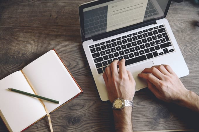 يدين لشخص ينقر على لوحة مفاتيح لابتوب و بجانبه كتاب و قلم 