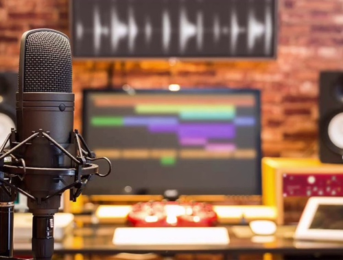 على يسار الصورة يظهر ميكروفون و في الخلفية جهاز كمبيوتر موضوع على طاولة