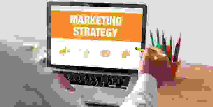 شخص يجلس الى طاولة امام حاسوبه ويمسك بيده قلم و تظهر على الشاشة عبارة MARKETING STRATEGY 