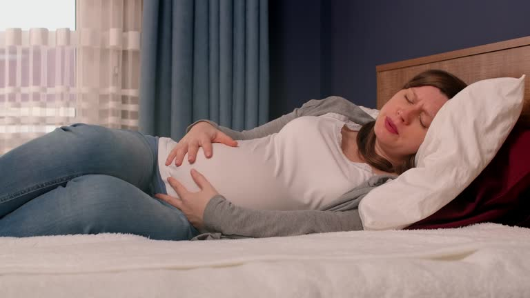 سيدة حامل مستلقية على السرير وهي تتألم و تضع يديها على بطنها 