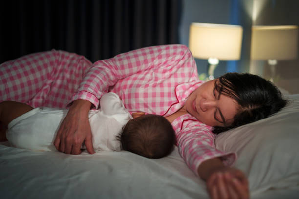 سيدة مستلقية على السرير ترتدي بيجامة زهرية و هي ترضع طفلها المستلقي بجانبها 