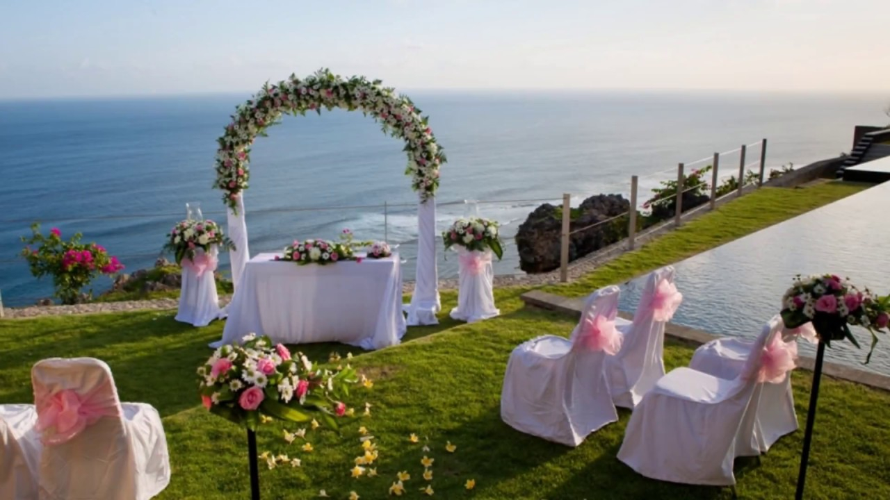 تحضير مكان الزفاف في مكان مغطى بالحشائش الخضراء مطل على البحر و تظهر طاولة العروسين و فوقهم اكليل من الزهور و كارسي الضيوف مزينة بالاغطية البيضاء و الورد الزهرية على مسند الكرسي من الخلف 