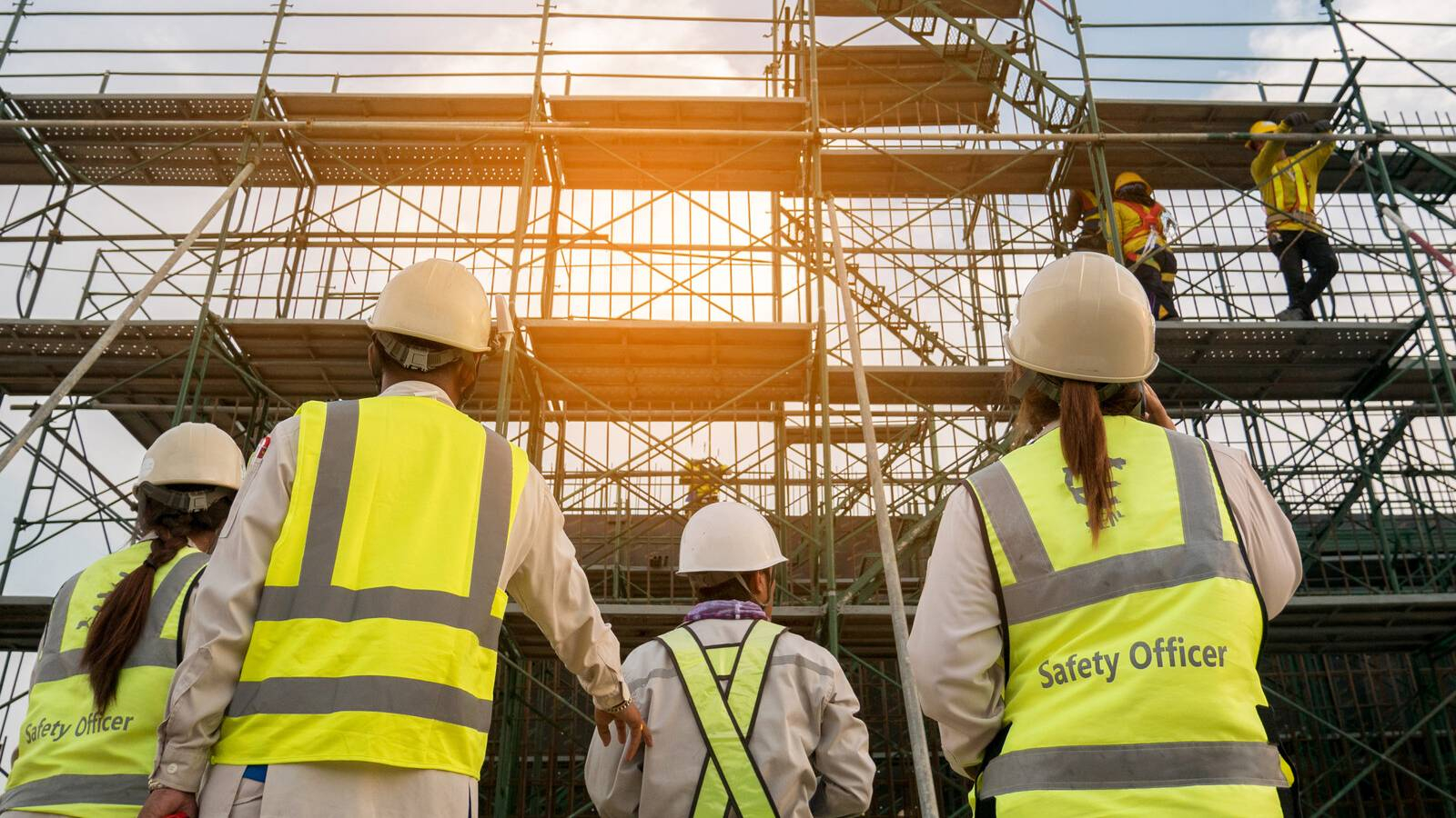 مجموعة من عمال البناء يعملون في البناء في مكان مرتفع وعلى الارض مجموعة من موظفين السلامة ويراقبون العمل و يقيمون المخاطر لتعزيز اجراءات السلامة 