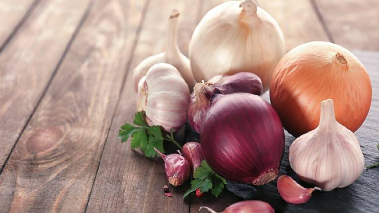 على طاولة خشبية يوجد حبات من البصل الاحمر والبرتقالي مع ثلاث حبات من الثوم و فصوص صغيرة بجانبها مع وريقات خضراء من البقدونس 