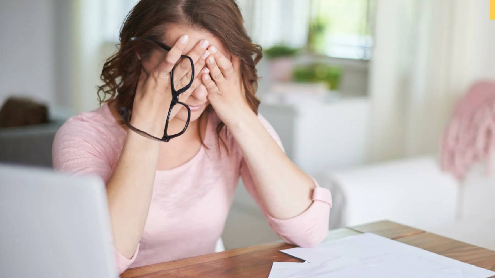 سيدة تضع يديها على وجهها وهي تحمل نظاراتها في اشارة الى التوتر وامامها اوراق عملها و جهاز كمبيوتر 