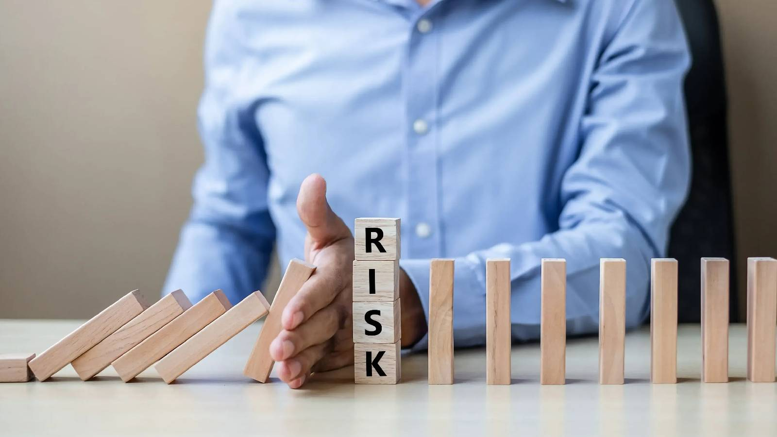 شخص يضع يده امام مجموعة من الالواح الخسبية و تظهر عبارة RISK على مكعبات خشبية و خلفها الواح خشبية تقف بتوازن 