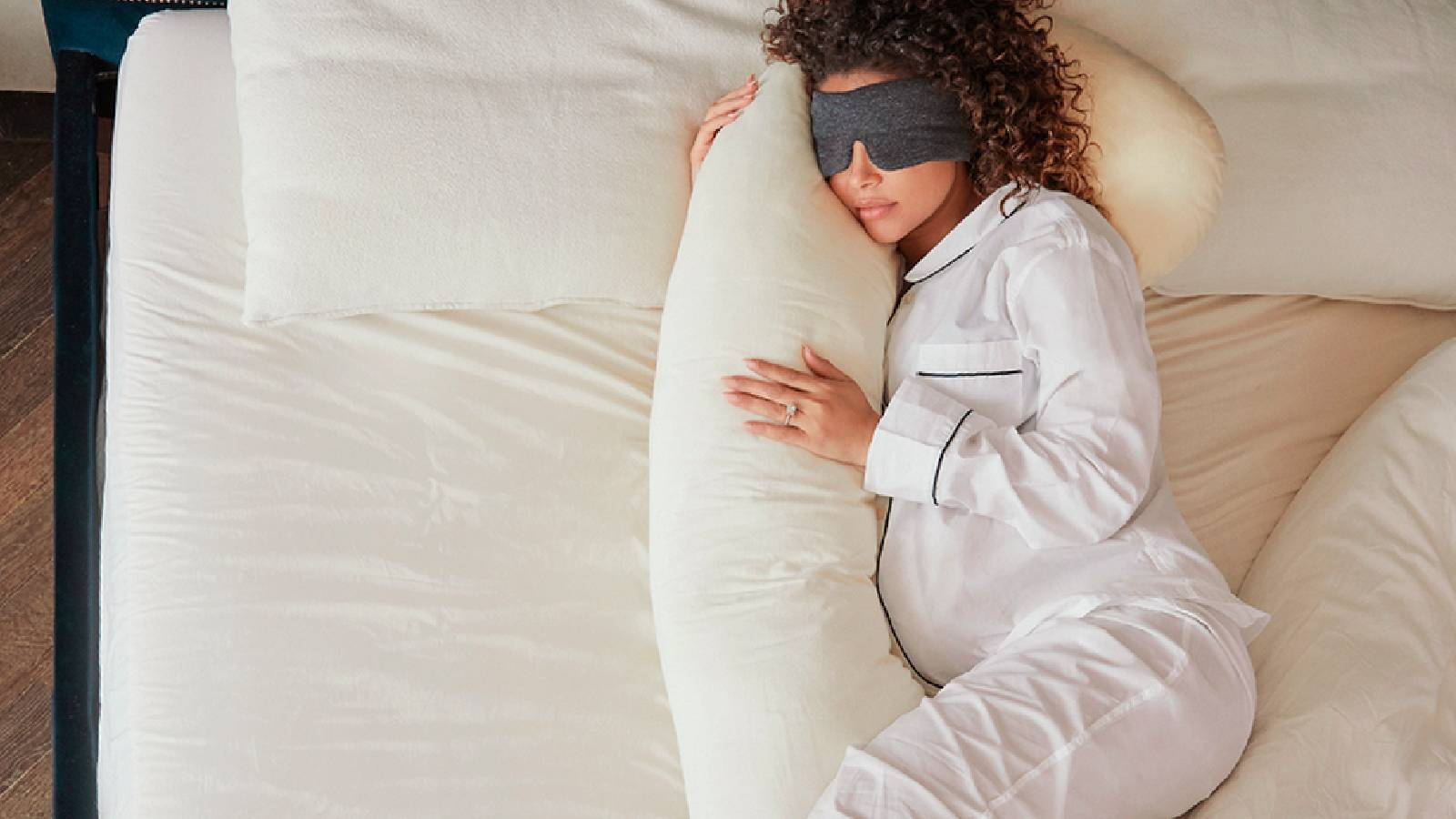 سيدة حامل تنام على سرير وتحتضن وسادة بيضاء وتضع على عيونها قطعة سوداء تساعدها على ابعاد الضوء عن عيونها 