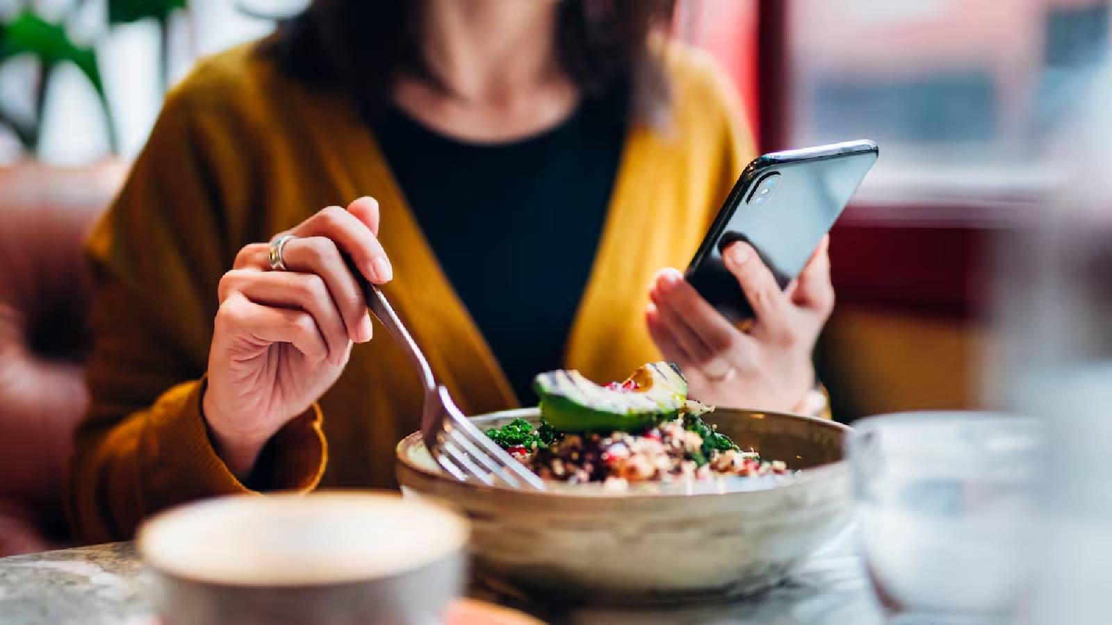 سيدة تجلس الى طاولة و في يدها اليسرى تحمل مويايل وتتصفحه و في يدها اليمنى تمسك بشوكه لتتناول طعامها 