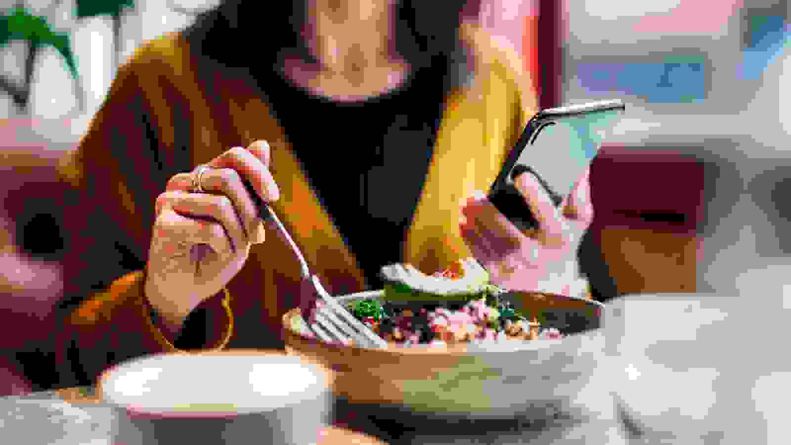 سيدة تجلس الى طاولة و في يدها اليسرى تحمل مويايل وتتصفحه و في يدها اليمنى تمسك بشوكه لتتناول طعامها 