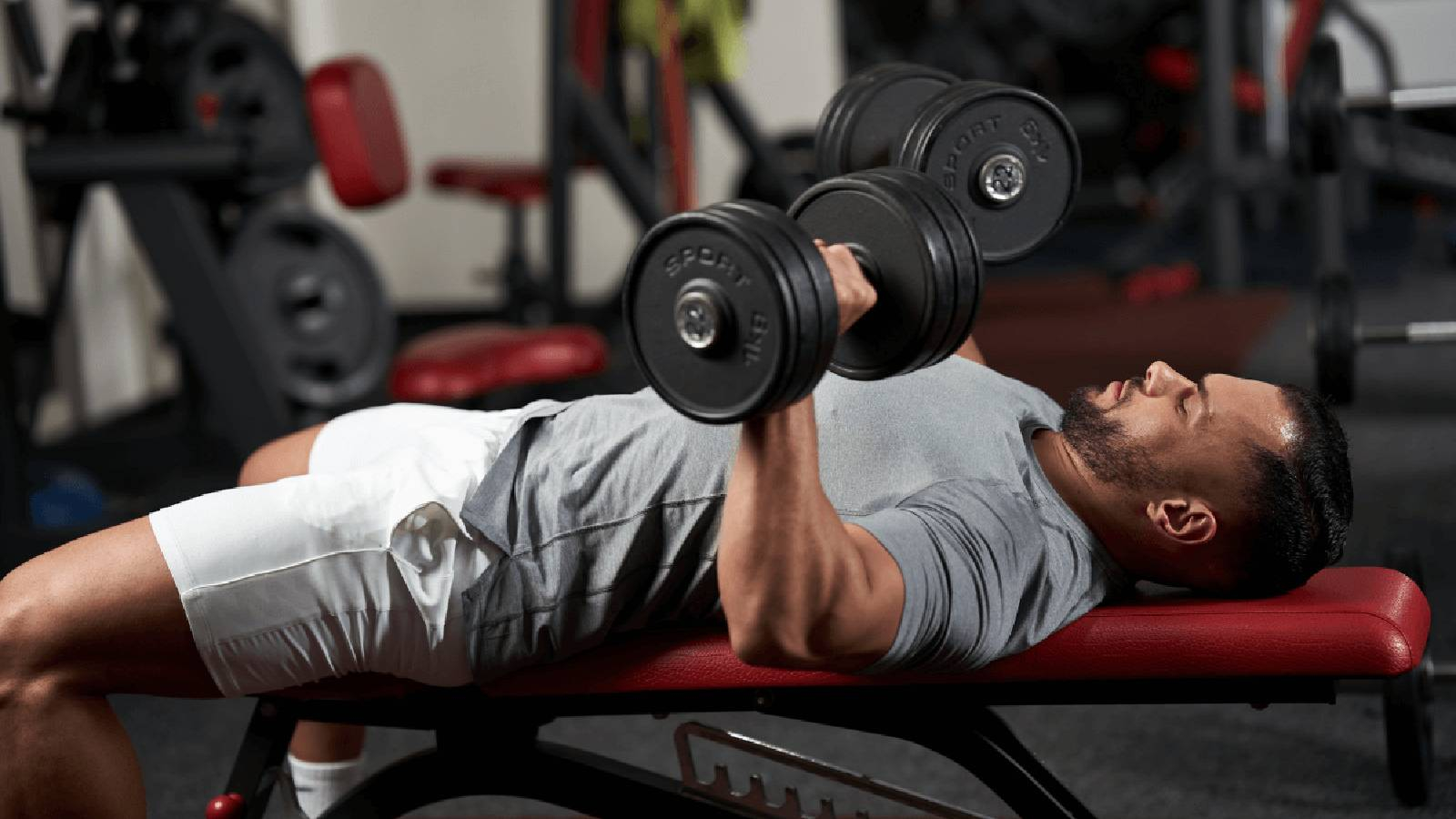رجل يرتدي تيشيرت رمادي و شورت ابيض مستبقي على طاولة التدريب الرفيعة و يحمل الاثقال بيديه ويرفعهم بالتوازي مع صدره 
