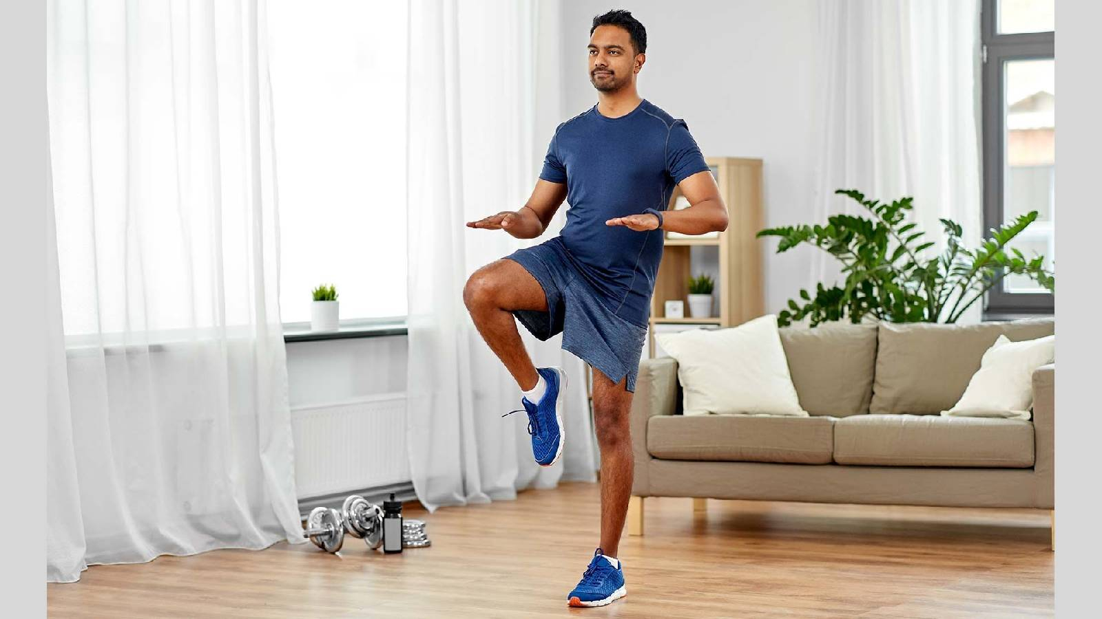 رجل يرتدي تيشيرت كحلي و شورت ازرق ويمارس الرياضة و هو يقف على رجل واحده  في غرفة الجلوس 