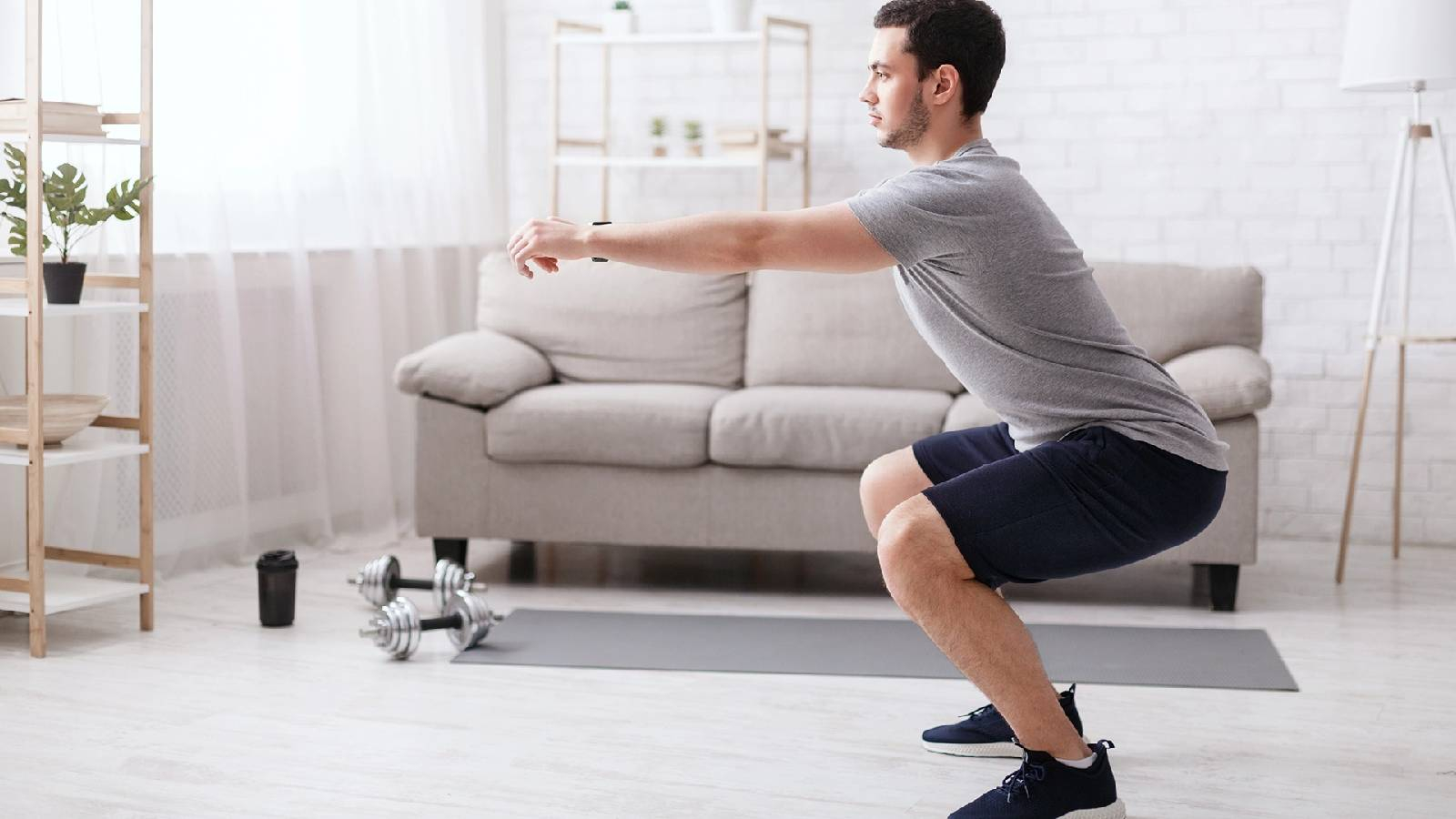 رجل يرتدي تيشيرت رمادي و شورت اسود يمارس تمارين السكوات آخذا وضعية الجلوس وهو مادا يديه للامام بشكل مستقيم 