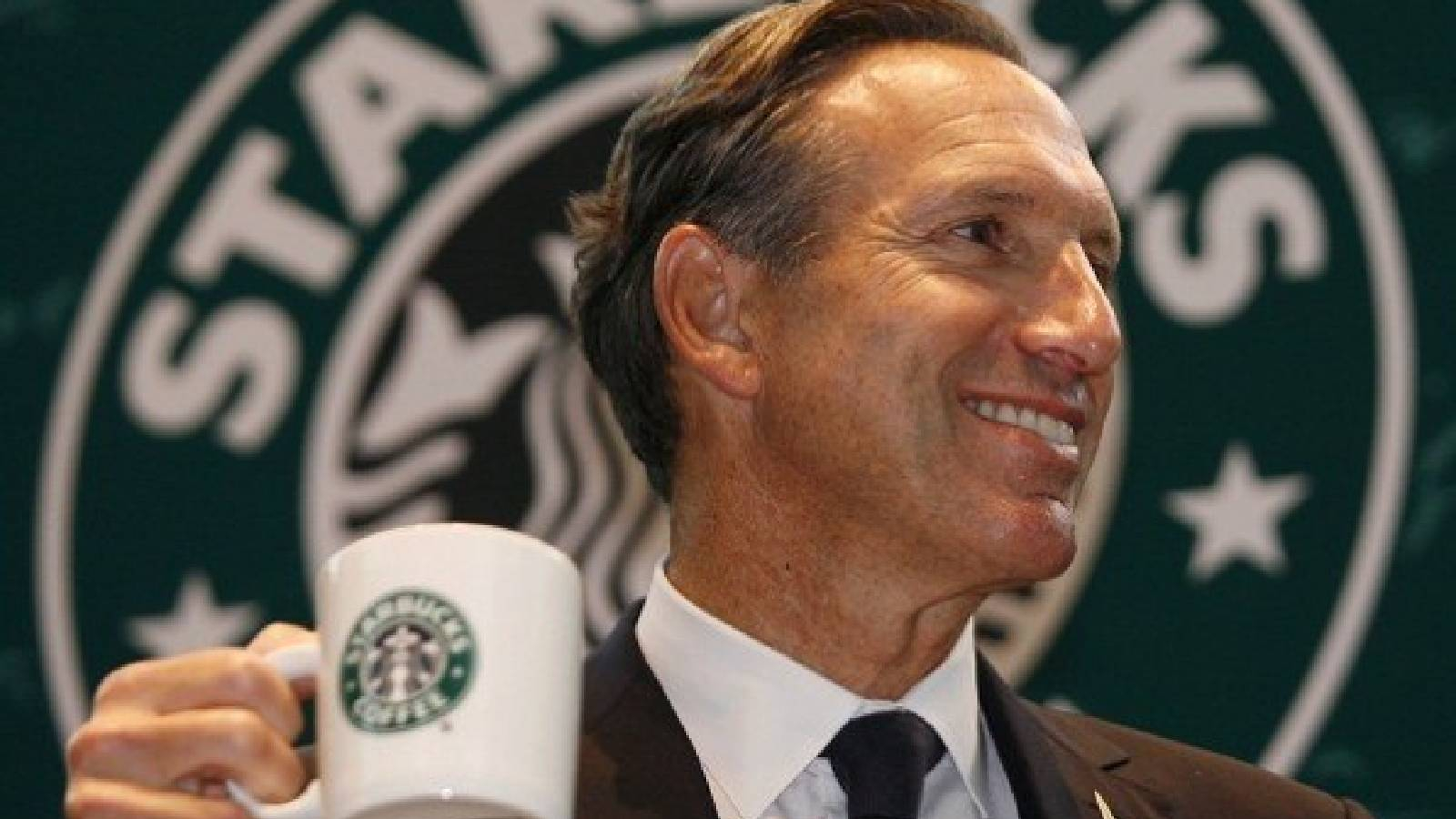 امام خلفية تحمل شعار ستاربكس يقف هوارد شولتتز يحمل كوبا ابيض من القهة مرسوم عليها شعار ستاربكس 