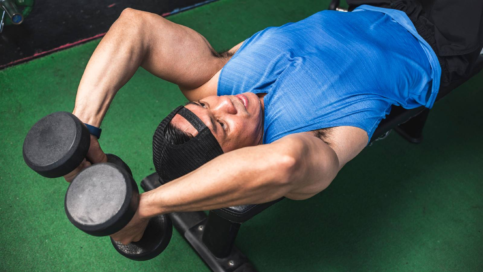 رجل النادي يرتدي تيشيرت ازرق وشورت اسود و هو مستلقي على طاولة التدريب و يحمل بيديه اثقال ويرفعها فوق رأسه هو مستلقي 