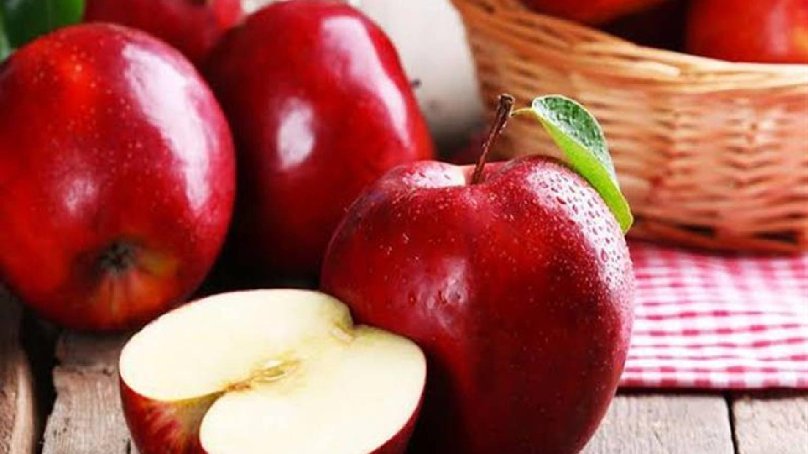 ثلاث ثمرات من التفاح الاحمر وبجانبهم نصف تفاحة  وعلى الجانب الايمن سلة من القش موضوعة على قطعة قماش من المربعات البيضاء والحمراء 