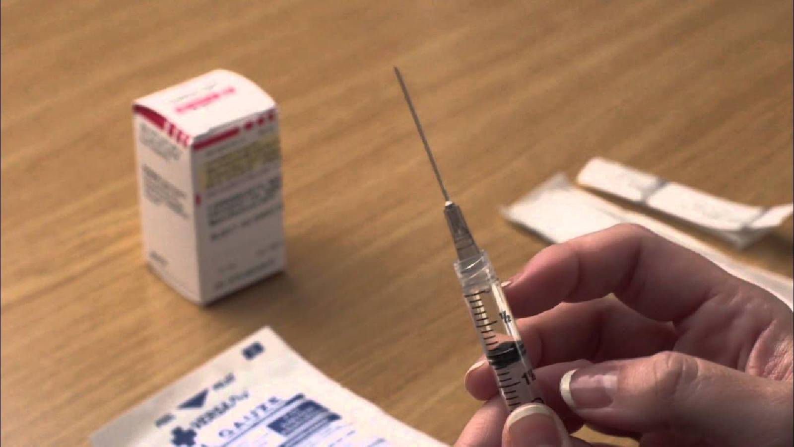 على طاولة بنية تظهر يد تحمل ابره مليئة بالدواء وامامها علبة دواء و