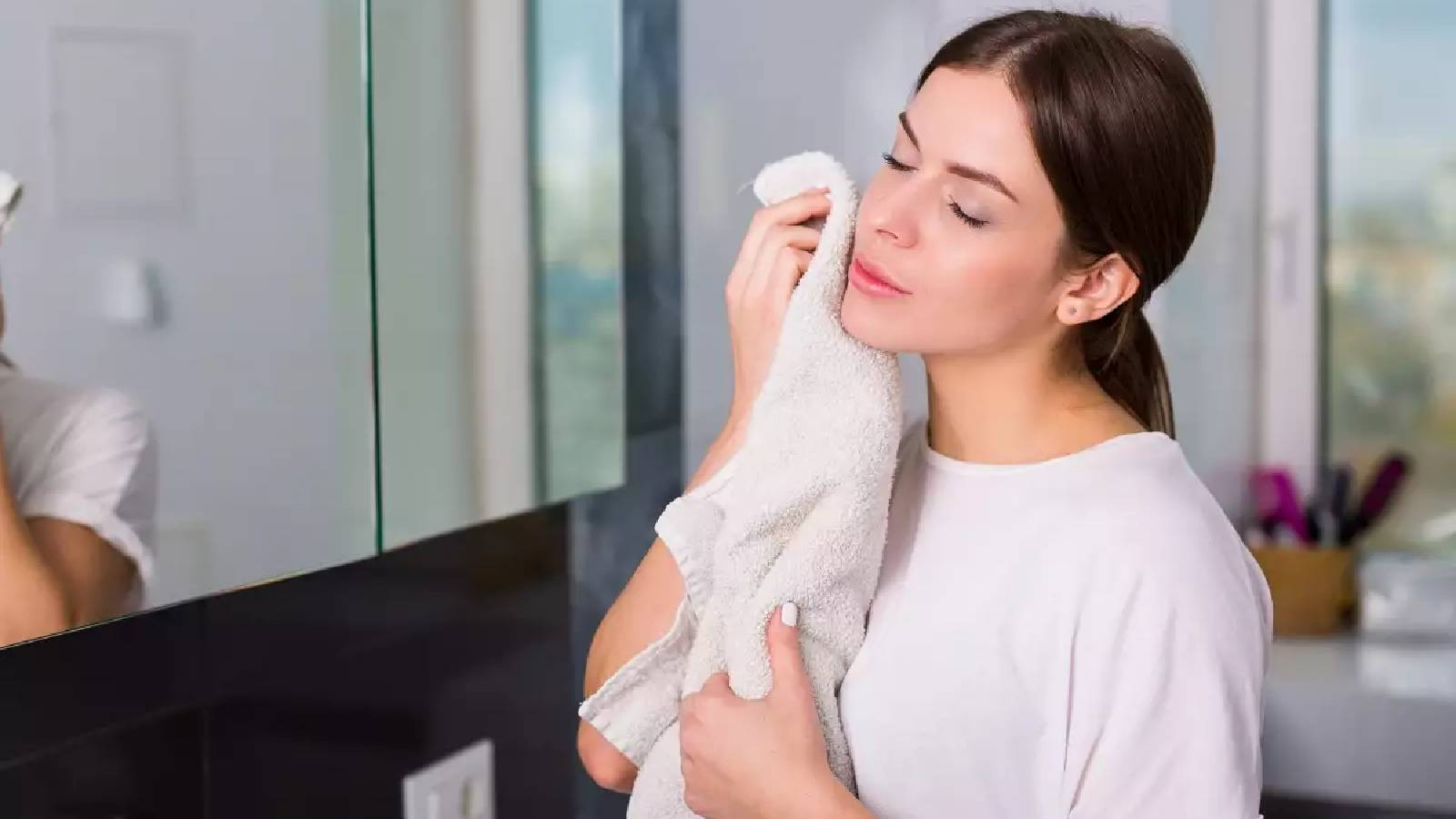 سيدة تحمل بيدها منشفة تجفف وجهها بعد تنظيفه وهي تقف امام المرآة 