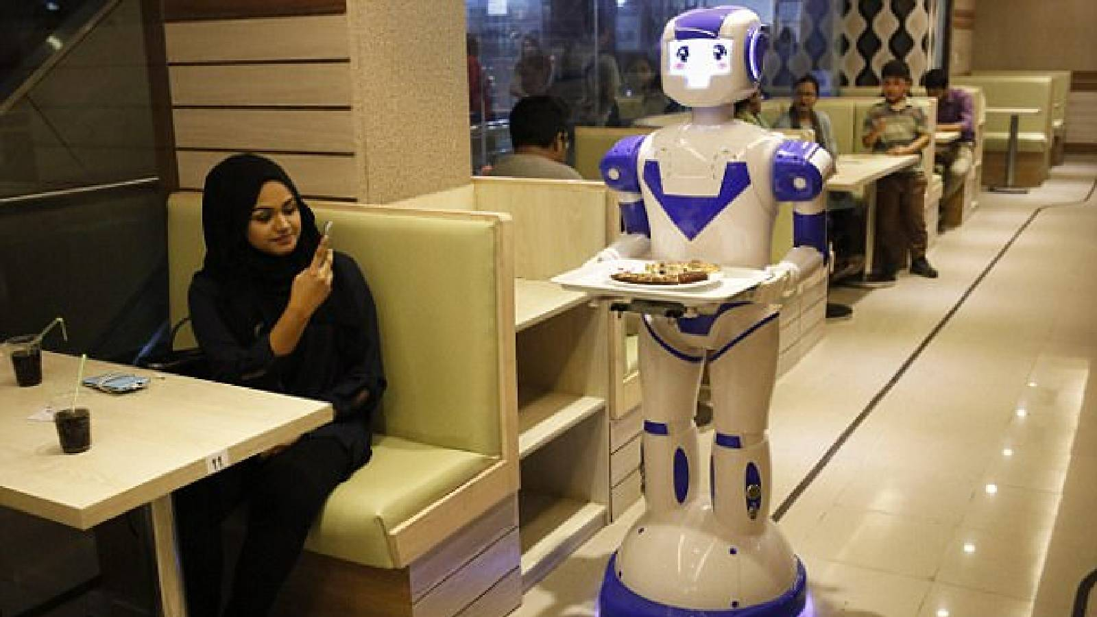 ضمن قاعة مطعم في الخليج تظهر سيدة وهي تلتقط صورة لروبوت آلي يقدم وجبة الطعام 
