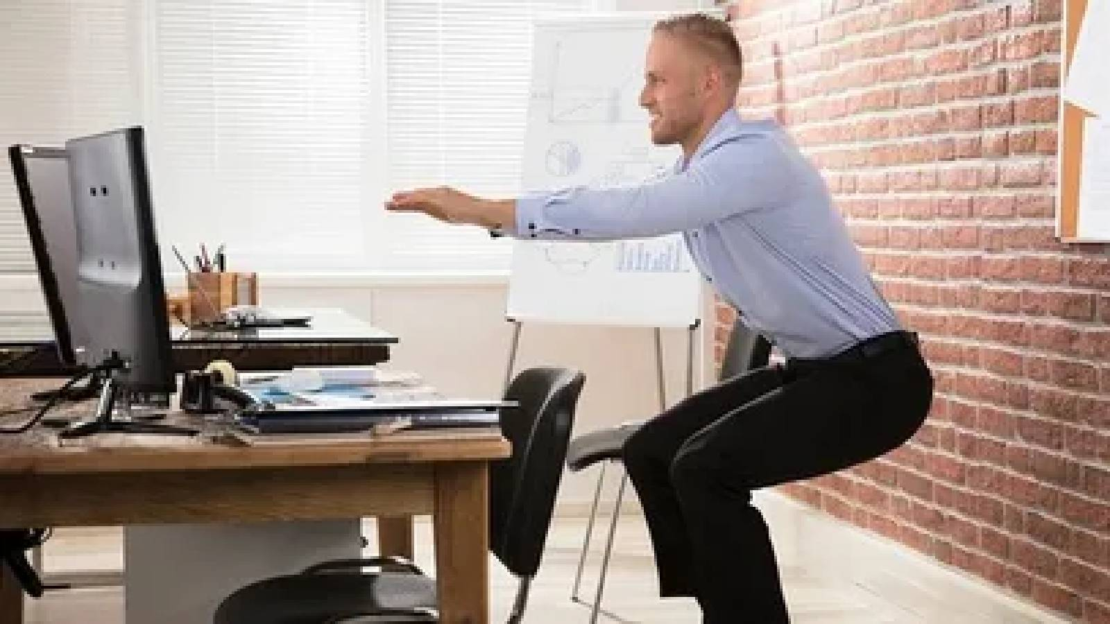 موظف يرتدي قميص ابيص وبنطلون اسود يقف خلف مكتبه يمارس تمرين الوقوف والجلوس 