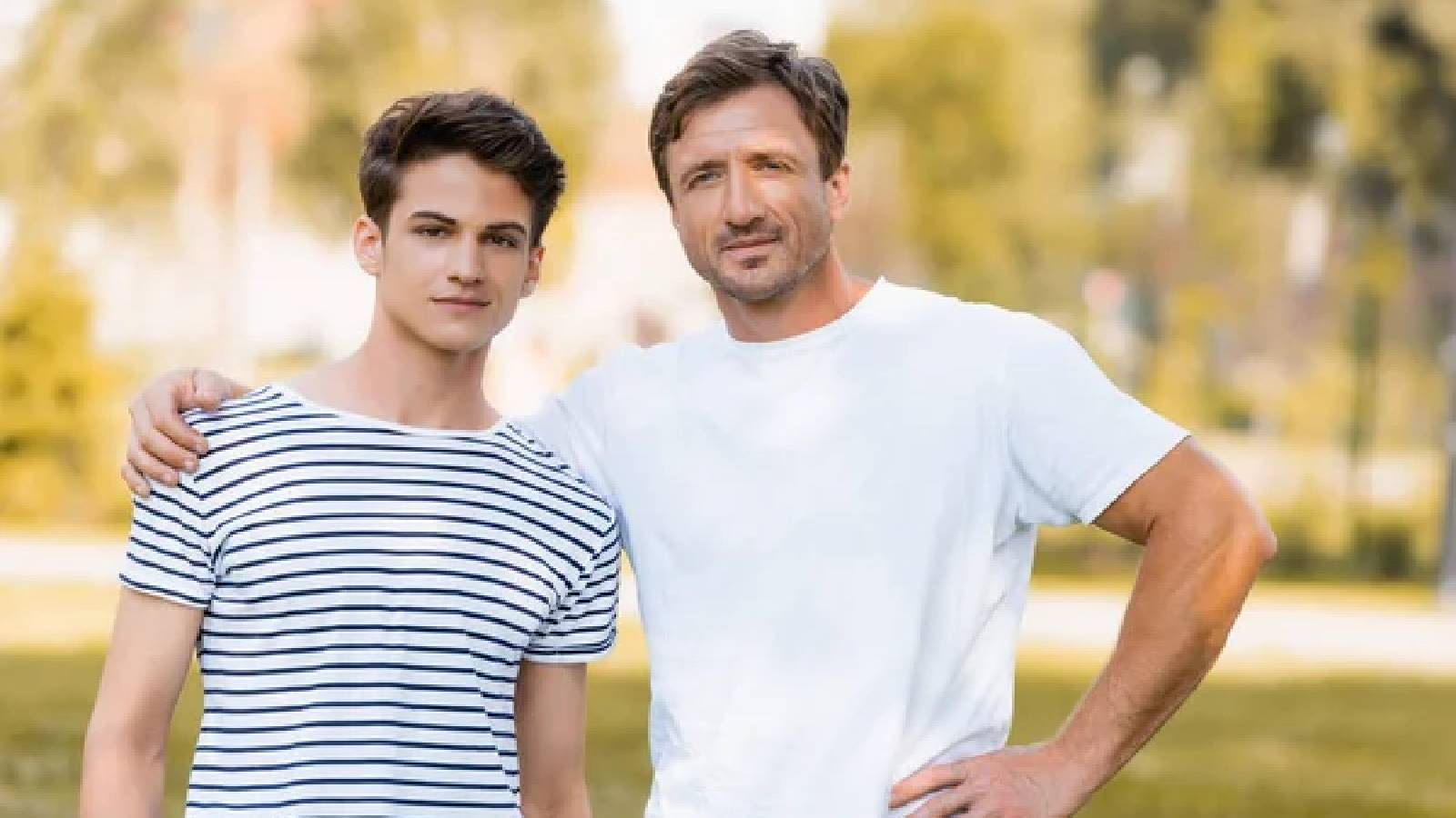 اب يرتدي تيشيرت ابيض يضع يده على كتف ابنه المراهق الذي يرتدي تيشيرت مخططة وهما يقفان في الهواء الطلق 