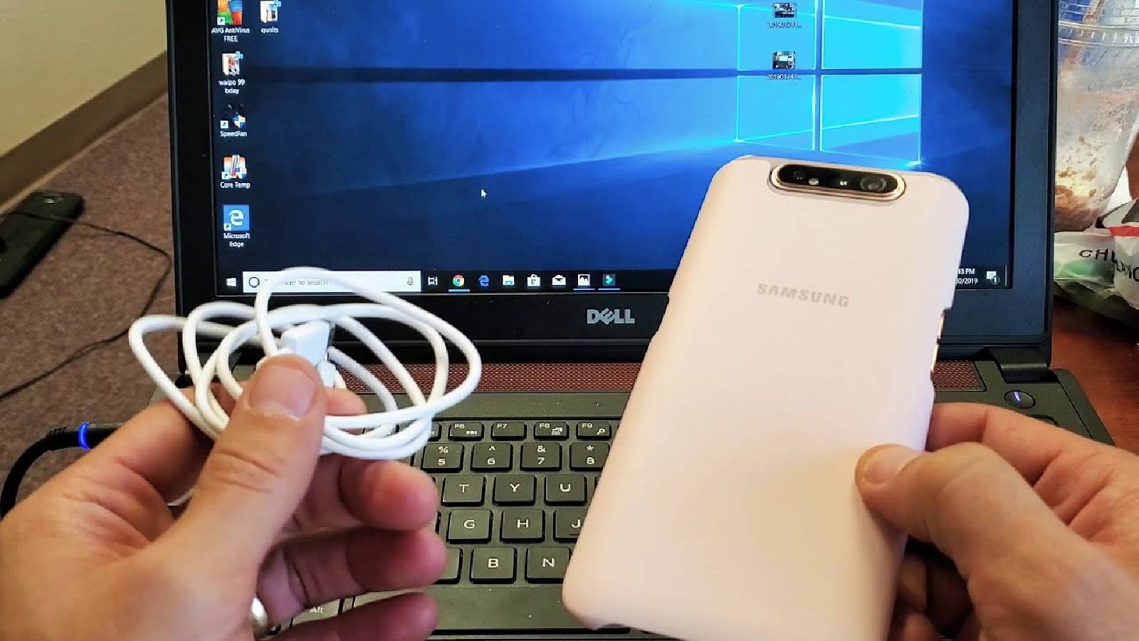 تظهر يدين الاولى تسمك جهاز سامسونغ ابيض والثاينة تمسك كابل sub امام جهاز لابتوب مفتوح 