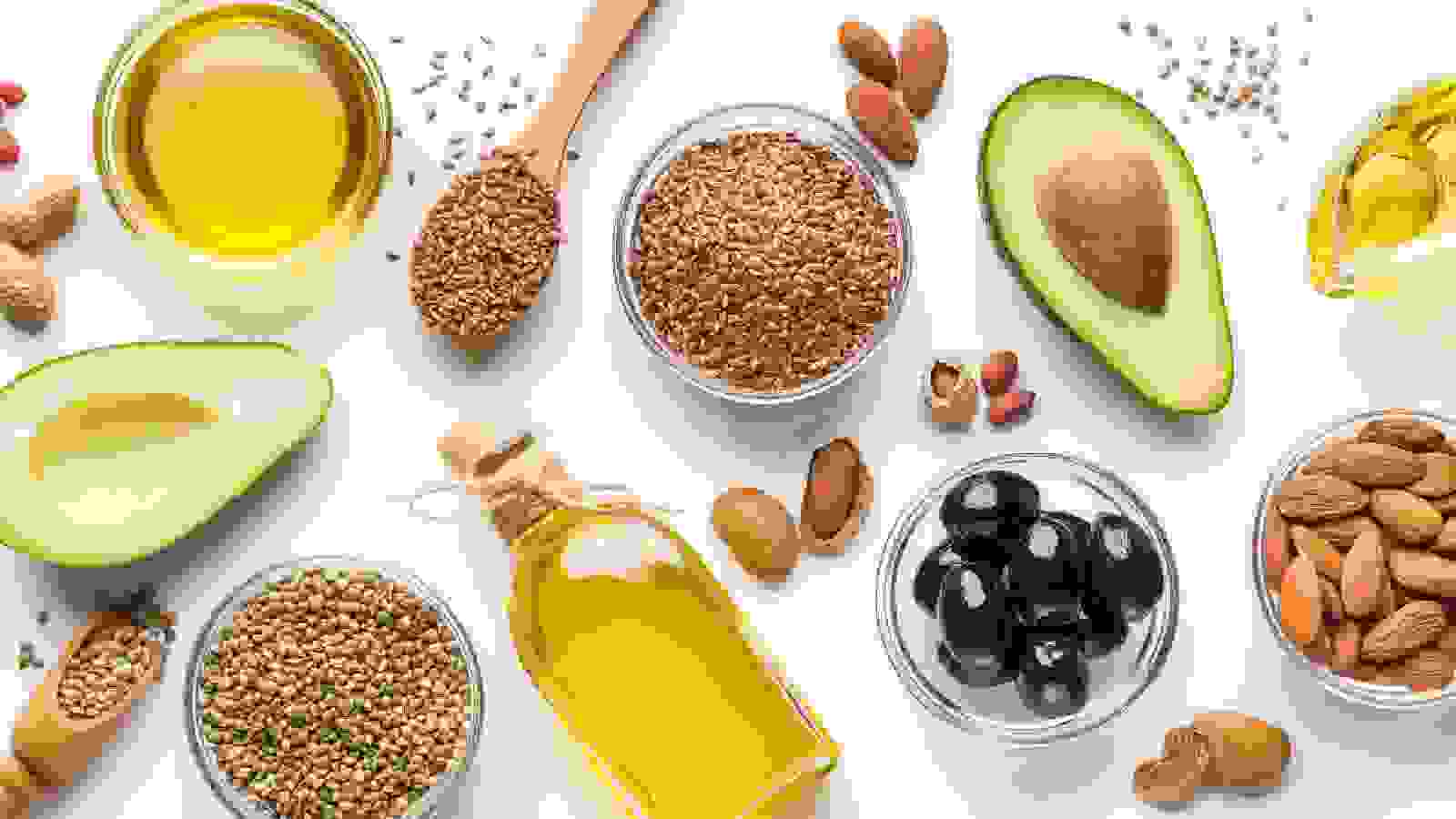 على خلفية بيضاء تظهر العديد من الاغذية الغنية بالدهون الصحية كزيت الزيتون و الافوكادو و اللوز وغيرها 