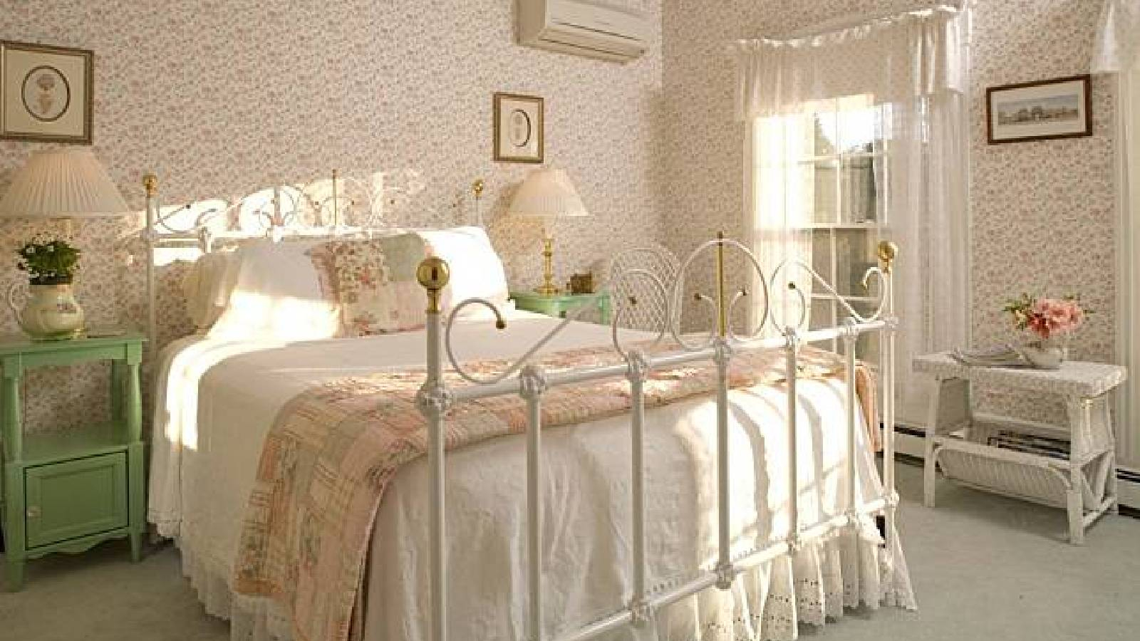 غرفة نوم ريفية بسرير ابيض من المعدن وعليه اغطية باللون الابيض والوردي وعلى الجهة اليمنى من الصورة يوجد نافذة صغيرة تزينها برادي بيضاء  وعلى جانبي السرير يوجد طاولتين باللون الاخضر افاتح وعليها مصباح انارة بالاضافة الى لوحات مطرزة باليد معلقة على الحيطان 