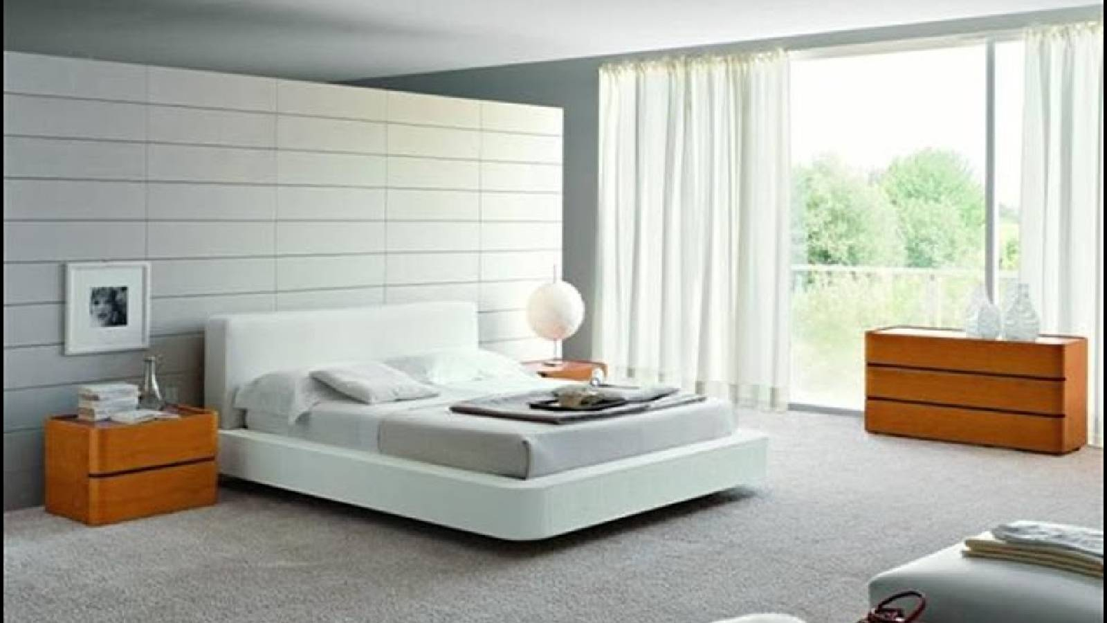 غرفة نوم حديثة تحوي سرير باللون الابيض وبجانبه طاولة خشبية وخلف السرير حائط ابيض و يوجد طاولة اكبر على الجهة اليمنى والغرفة تطل على بلكونة وواجهة زجاجية كبيرة عليها برادي بيضاء 