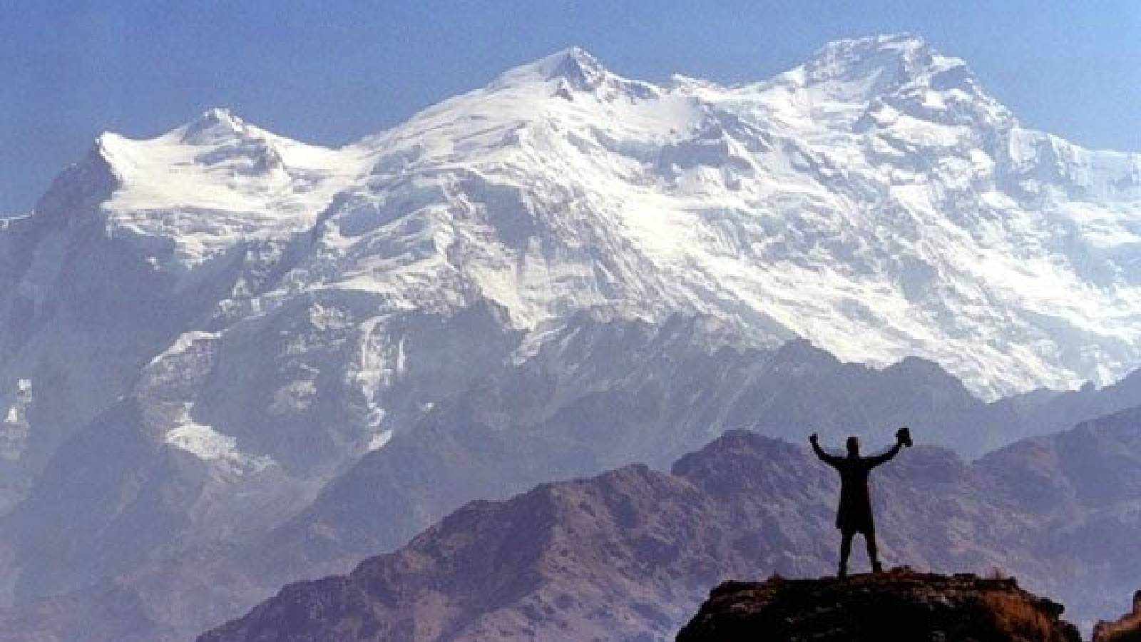 قمة جبلية عالية وضخمة مغطاة بالثلوج البيضاء و يقف على شفح جبل مقالبل له رجل رافع يديه 