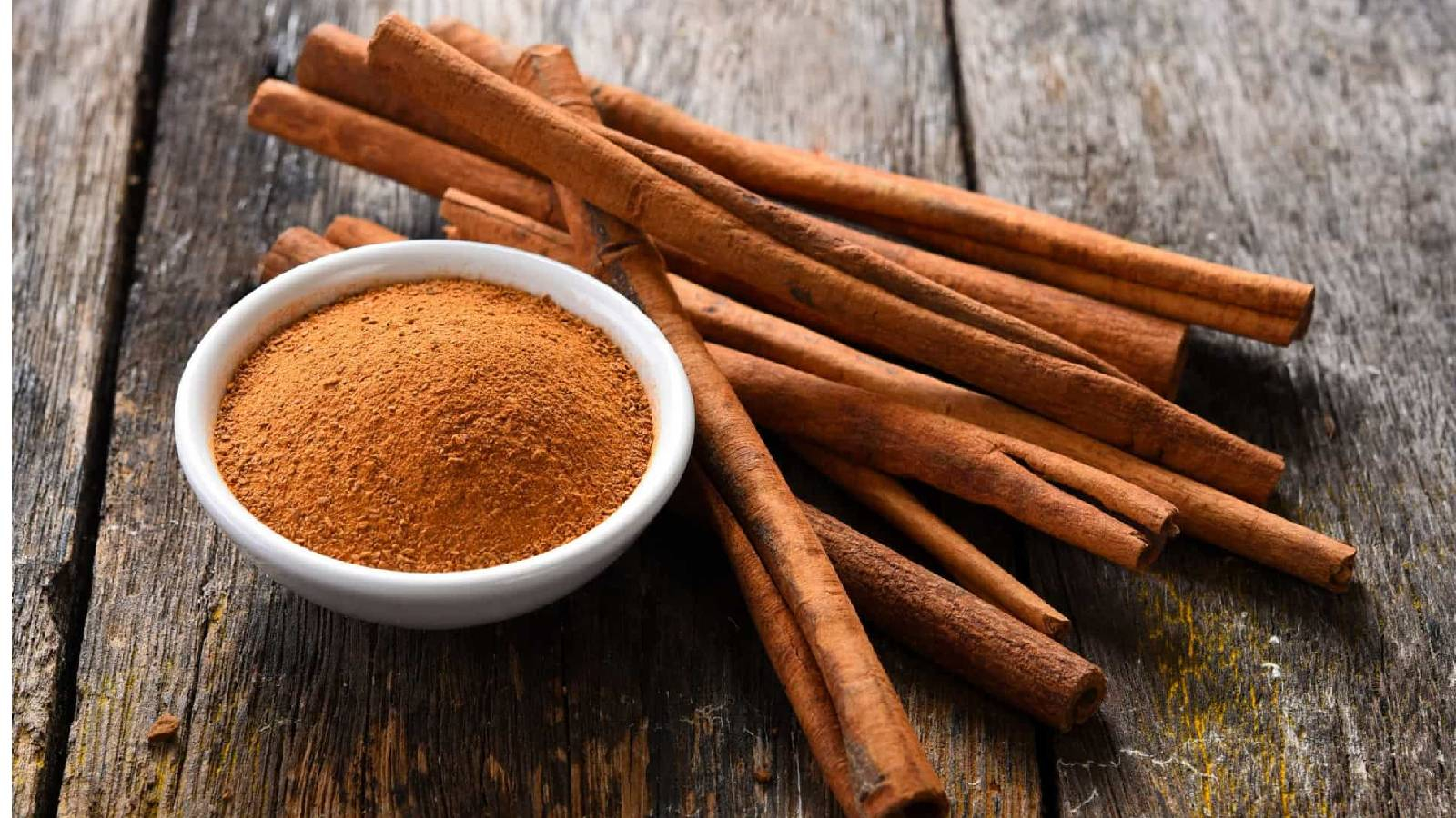 على طاولة خشبية يوجد مجموعة من اعواد القرفة وبجانبها طبق ابيض يحوي مطحون القرفة 